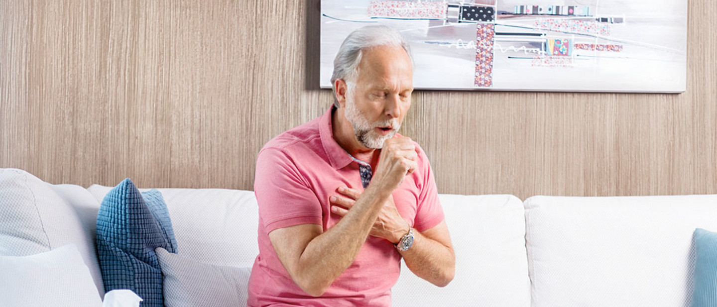 Mucociliary clearance Older man sitting on a sofa in a bright living room with one hand on his chest and coughing into the other.