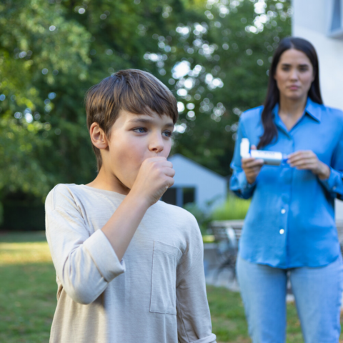 My child has asthma and is ill all the time – tips from the paediatrician Emmanuel Nellen Junge hustet und hält sich die Faust vor dem Mund, im Hintergrund ist Mutter mit Asthmaspray zu sehen
