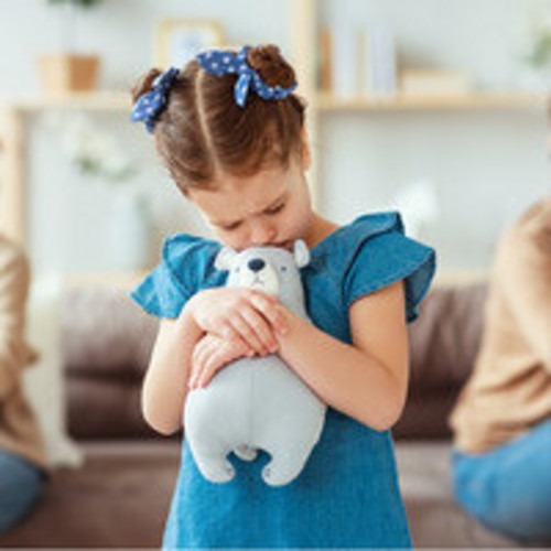 Ein junges Mädchen in einem blauen Kleid mit blauen Schleifen im Haar hält traurig ein Stofftier in den Armen. Im Hintergrund sitzen ihre Eltern auf einem Sofa, mit verschränkten Armen und abgewandten Blicken, scheinbar in einem Streit. Die Szene vermittelt ein Gefühl von Traurigkeit und Unsicherheit.