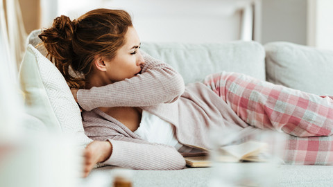 Warum habe ich Reizhusten im Liegen? Antworten von Prof. Dr. Fischer Junge Frau liegt auf der Couch und hustet in ihre Armbeuge