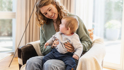 Infections in newborn infants & toddlers – what can you do if they have sniffles or a cough? Interview with Dr Manfred Praun Junge Mutter sitzt mit Baby auf dem Schoß in einem Sessel