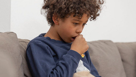 Chronic cough – interview with lung specialist Prof. Dr Rainald Fischer Junge sitzt auf dem Sofa und hält sich beim Husten eine Faust vor den Mund