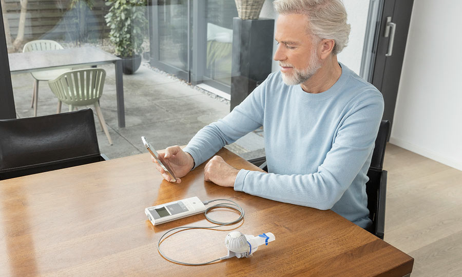 Ein älterer Mann sitzt an einem Holztisch in einem Wohnraum. Vor ihm liegt ein eFlow®rapid Inhalationssystem. Er hält ein Smartphone in der Hand und schaut konzentriert darauf.