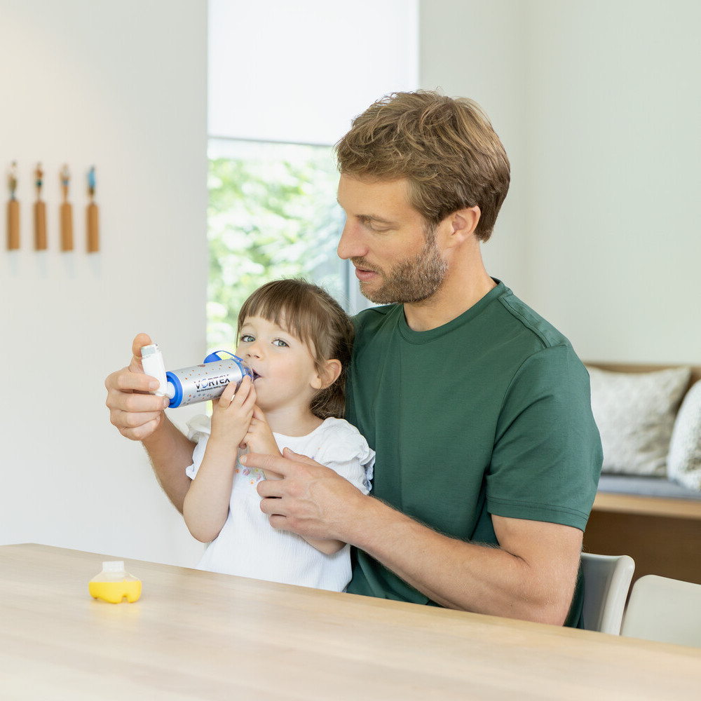 Vater sitzt mit Tochter am Tisch und hilft ihr dabei Dosieraerosol mit Spacer zu inhalieren