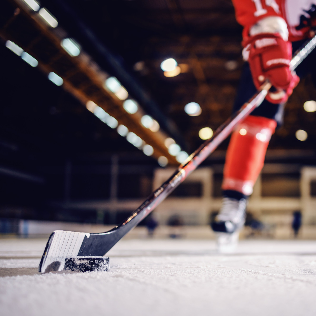 Nahaufnahme eines Hockeyschlägers mit Puk auf dem Eis, im Hintergrund sind Beine des Spielers zu erkennen
