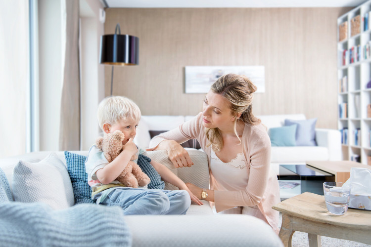 Tipps bei Erkältung beim Kind: Hals vom Schleim befreien Kleines Kind sitzt auf der Couch, Mutter kniet neben ihm und redet ihm gut zu