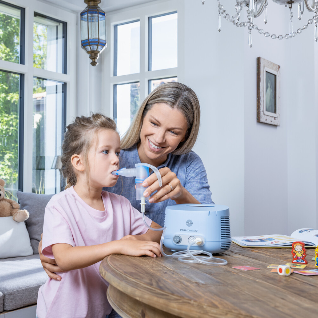 Junge Mutter sitzt mit Tocher am Tisch und hilft ihr beim Inhalieren mit einem PARI COMPACT2 Inhalationsgerät