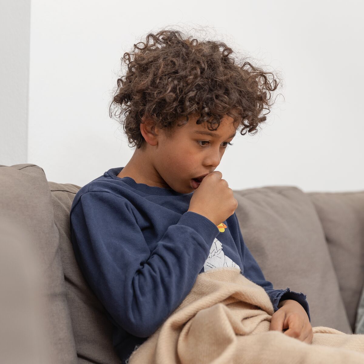 Junge sitzt auf dem Sofa und hält sich beim Husten eine Faust vor den Mund