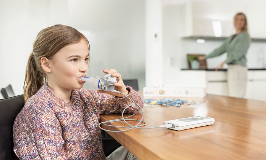 Ein junges Mädchen inhaliert mit dem eflow rapid und eTrack zu Hause. Die Mutter steht im Hintergrund in der Küche und beobachtet die Tochter dabei