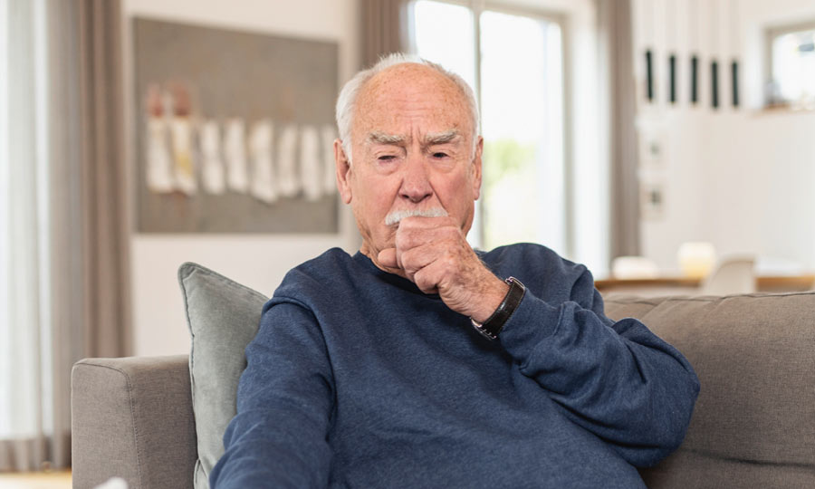 Ein älterer Mann mit weißen Haaren und Schnurrbart sitzt auf einem grauen Sofa in einem modern eingerichteten Wohnzimmer. Er trägt einen dunkelblauen Pullover und hat eine nachdenkliche Miene, während er in seine Hand hustet. Im Hintergrund sind große Fenster mit Vorhängen, eine abstrakte Wanddekoration und ein Essbereich mit Stühlen zu sehen.