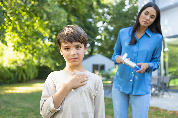 Ein Junge mit Asthma hält sich die Hand an die Brust, im Hintergrund steht sorgenvoll seine Mutter