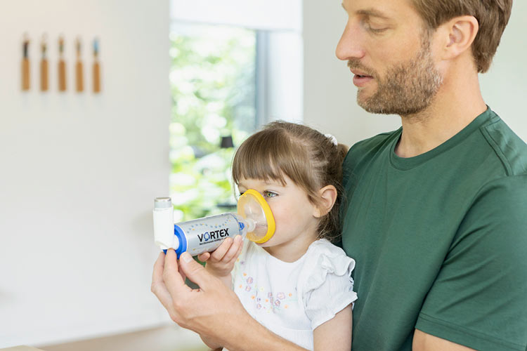 Ein kleines Mädchen inhaliert ein Asthmaspray mit der VORTEX Inhalierhilfe und Maske. Ihr Vater hilft ihr dabei