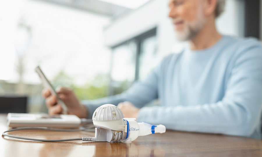 eFlow rapid und eTrack Controller liegen auf einem Tisch im Wohnzimmer. Ein Mann im Hintergrund kontrolliert mit seinem Handy und der PARI Connect App das Inhalationsergebnis