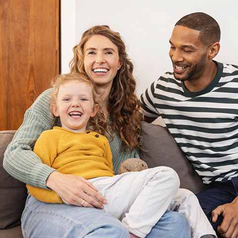 Eine lachende Familie sitzt gemeinsam auf einem Sofa. Die Mutter mit langen, welligen Haaren trägt einen grünen Pullover und hält ihre junge Tochter auf dem Schoß, die fröhlich in die Kamera lacht und einen gelben Pullover trägt. Der Vater, mit kurzem Bart und gestreiftem T-Shirt, sitzt daneben und lächelt die beiden an. Die Szene wirkt warm und harmonisch.