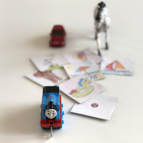 Eine kleine blaue Spielzeuglokomotive steht im Vordergrund auf einem weißen Tisch. Dahinter liegen mehrere handgezeichnete Karten mit bunten Motiven wie Rutschen und Booten. Im Hintergrund sind ein rotes Spielzeugauto und eine kleine Zebrafigur zu sehen. Die Szene vermittelt ein spielerisches Reise- oder Abenteuermotiv.