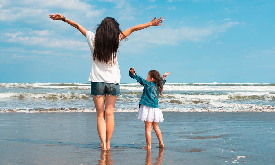 Eine Frau mit langen dunklen Haaren und ein kleines Mädchen stehen barfuß am Strand mit ausgebreiteten Armen, während die Wellen des Meeres an den Sand rollen. Der Himmel ist blau mit wenigen Wolken.