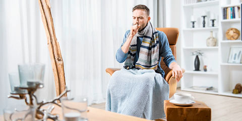 Ein junger Mann mit Schal und Decke sitzt in einem gemütlichen Wohnzimmer auf einem Sessel und hustet. Vor ihm steht ein Tisch mit einer Tasse Tee.