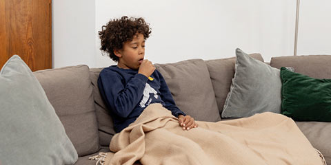 Ein Junge mit lockigen Haaren sitzt krank auf einem grauen Sofa, eingewickelt in eine beige Decke. Er hustet in seine Hand.