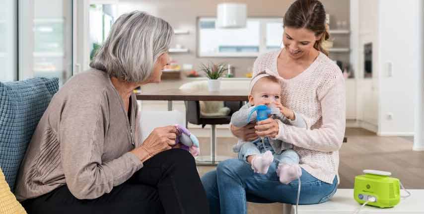 Eine junge Mutter sitzt mit ihrem Baby auf dem Schoß in einem modernen Wohnzimmer. Sie hilft dem Baby, eine blaue Inhalationsmaske zu benutzen. Eine ältere Frau mit grauen Haaren, vermutlich die Großmutter, sitzt daneben auf einem Sofa und hält ein Kuscheltier, während sie die beiden liebevoll ansieht. Ein PARI BOY Junior Inhalationsgerät steht auf dem Tisch.