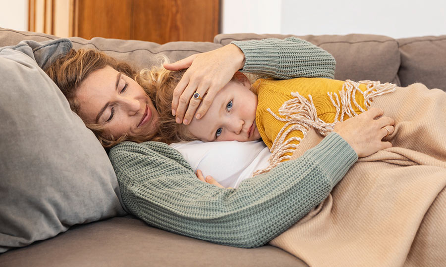 Mother holds her child's forehead and lies with him on the sofa