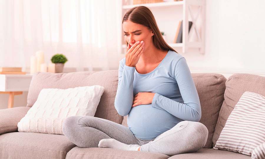 A pregnant woman sits on the sofa with a cold and holds her hand in front of her mouth