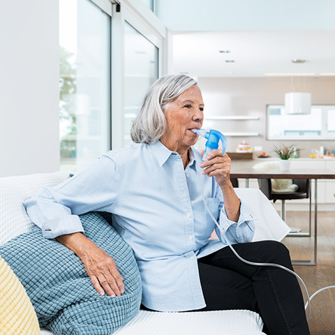 Elderly woman sitting on a couch using a nebulizer device for respiratory treatment in a bright, modern living room.