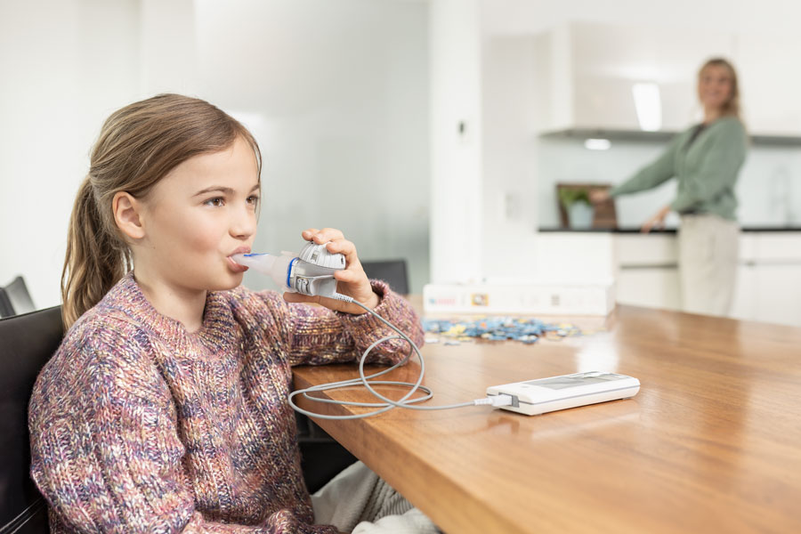 A girl sits on a chair in front of a table and inhales with the eFlow®rapid.