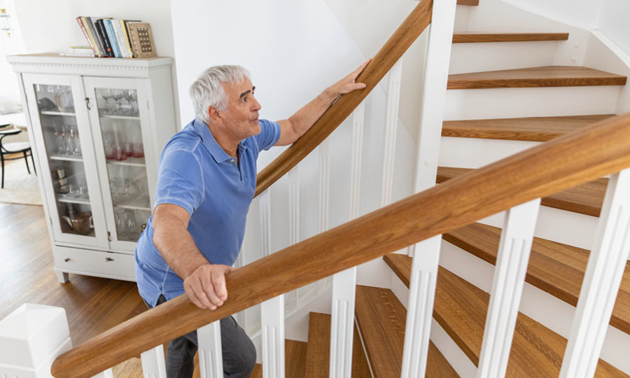 Climbing stairs can become a real challenge with COPD An elderly man slowly climbs a wooden staircase. He holds onto the railing with both hands and appears exhausted or short of breath.